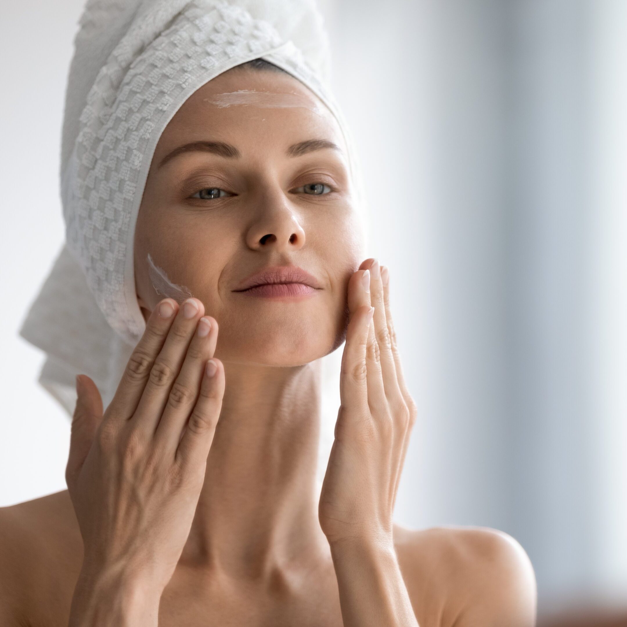 Closeup head shot pleasant beautiful woman applying moisturizing creme on face after shower. Smiling young pretty lady wrapped in towel smoothing perfecting skin, daily morning routine concept.
