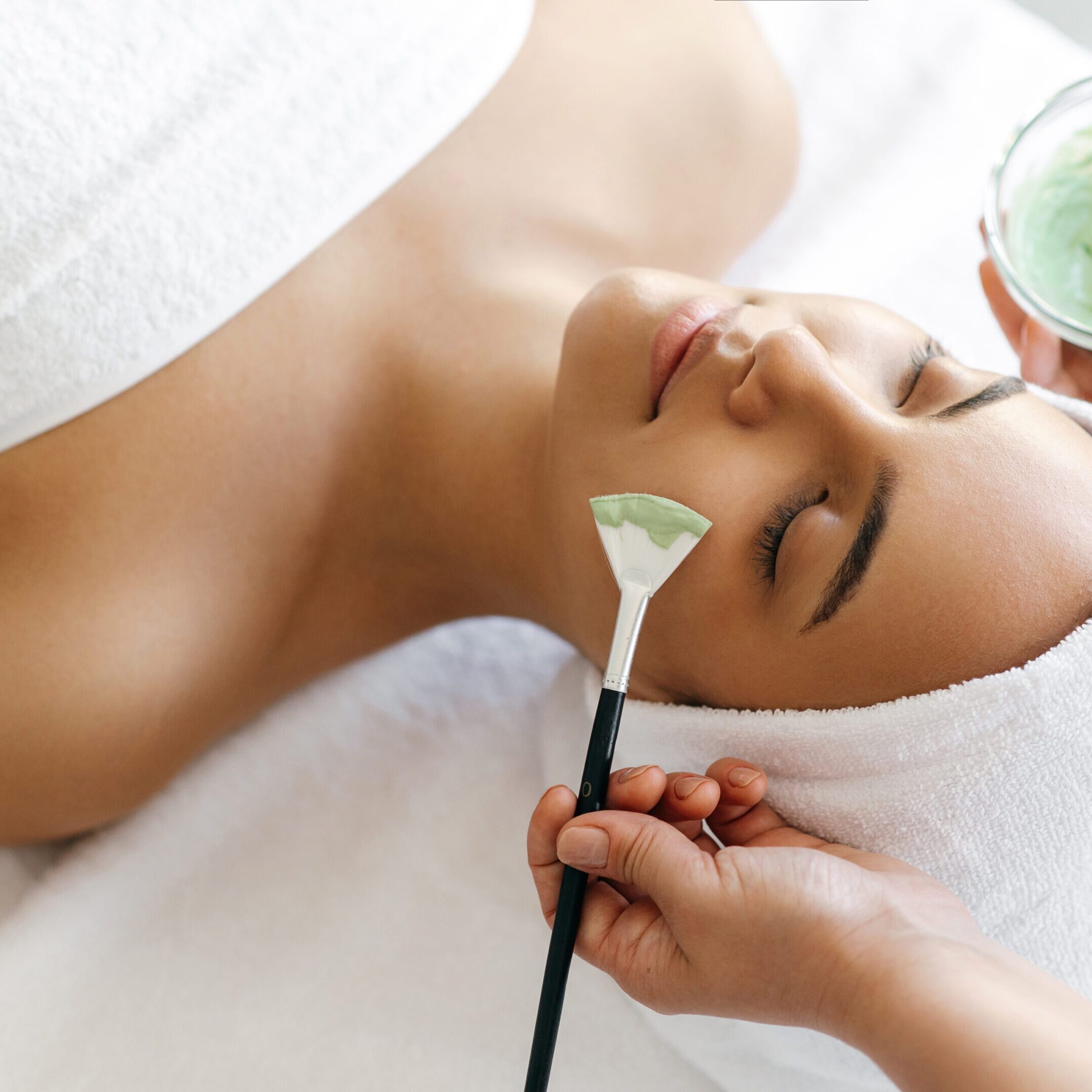 Facial treatments, wrinkle prevention, facial cleansing. Top view of beauty procedure, therapist applying green face mask on the face of a beautiful brazilian or hispanic young woman